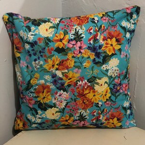 May include: Square cushion with a bright floral design. The cushion has a light blue base, covered in colourful flowers in yellow, orange, pink, and purple. The cushion is square and seems to be made of fabric.