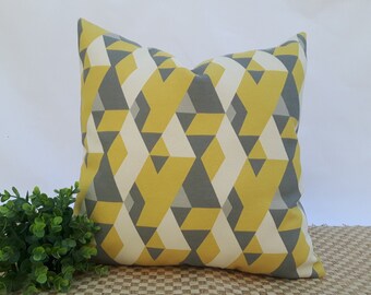 May include: Square decorative pillow with a geometric pattern in shades of yellow, gray, and off-white. The pillow features a repeating design of angled shapes, creating a modern and stylish look. The pillow is placed on a woven surface.