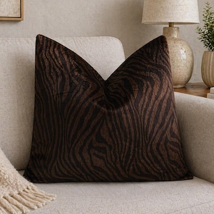 Könnte beinhalten: Ein quadratisches Zierkissen mit einem braunen und schwarzen Zebra-Streifenmuster. Das Kissen hat eine weiche, strukturierte Oberfläche und liegt auf einem hellen Sofa. Der Animal-Print verleiht der Einrichtung einen Hauch von Wildnis.