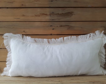 May include: White rectangular pillow with a white fringe border. The pillow is sitting on a wooden surface.