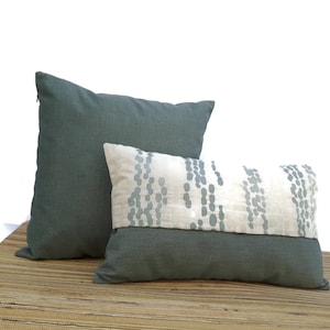 Op de afbeelding: Twee decoratieve sierkussens. Eén is een effen vierkant kussen in de kleur teal. De andere is een rechthoekig kussen met een tealkleurige onderkant, een witte midden met een teal stippenpatroon en een tealkleurige bovenkant. Beide kussens liggen op een geweven oppervlak.