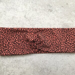 Könnte beinhalten: Ein korallenfarbenes Stirnband mit einem schwarzen Leopardenmuster. Der Stoff ist oben verdreht, wodurch ein stilvolles Accessoire entsteht. Dieses Stirnband ist ein modisches Haaraccessoire.