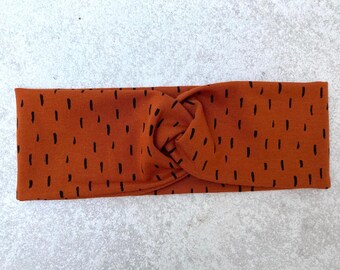 May include: A brown headband with a black abstract pattern. The headband is twisted and knotted in the center.