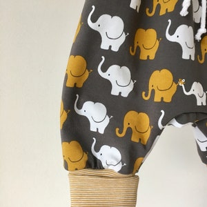 K&ouml;nnte beinhalten: Graue Babyhose mit einem Muster aus wei&szlig;en und gelben Elefanten. Die Hose hat einen braunen und wei&szlig;en gestreiften Bund.