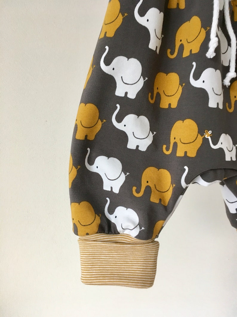 K&ouml;nnte beinhalten: Graue Babyhose mit einem Muster aus wei&szlig;en und gelben Elefanten. Die Hose hat einen braunen und wei&szlig;en gestreiften Bund.