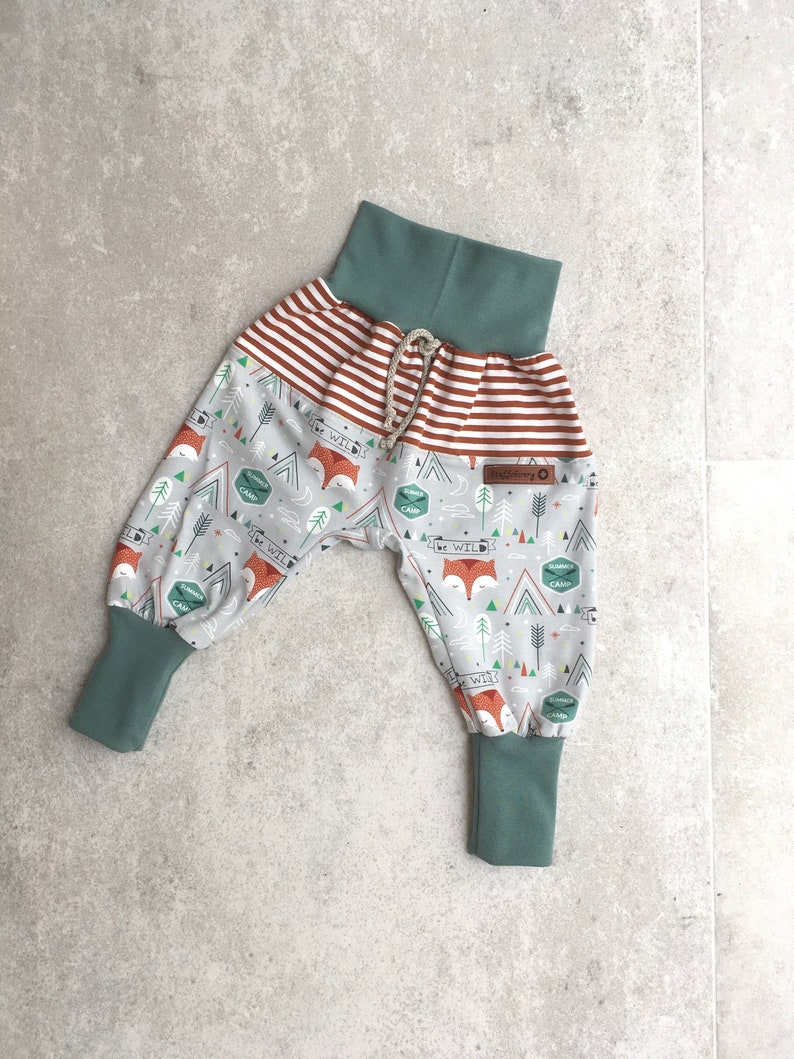May include: Gray and white patterned pants with a red and white striped waistband. The pants have a drawstring and green cuffs. The pattern features foxes, trees, and the words "Be Wild" and "Summer Camp".