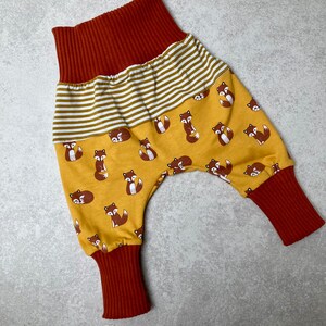 May include: Mustard yellow baby pants with a fox print and red ribbed cuffs. The pants have a white and mustard yellow striped waistband.