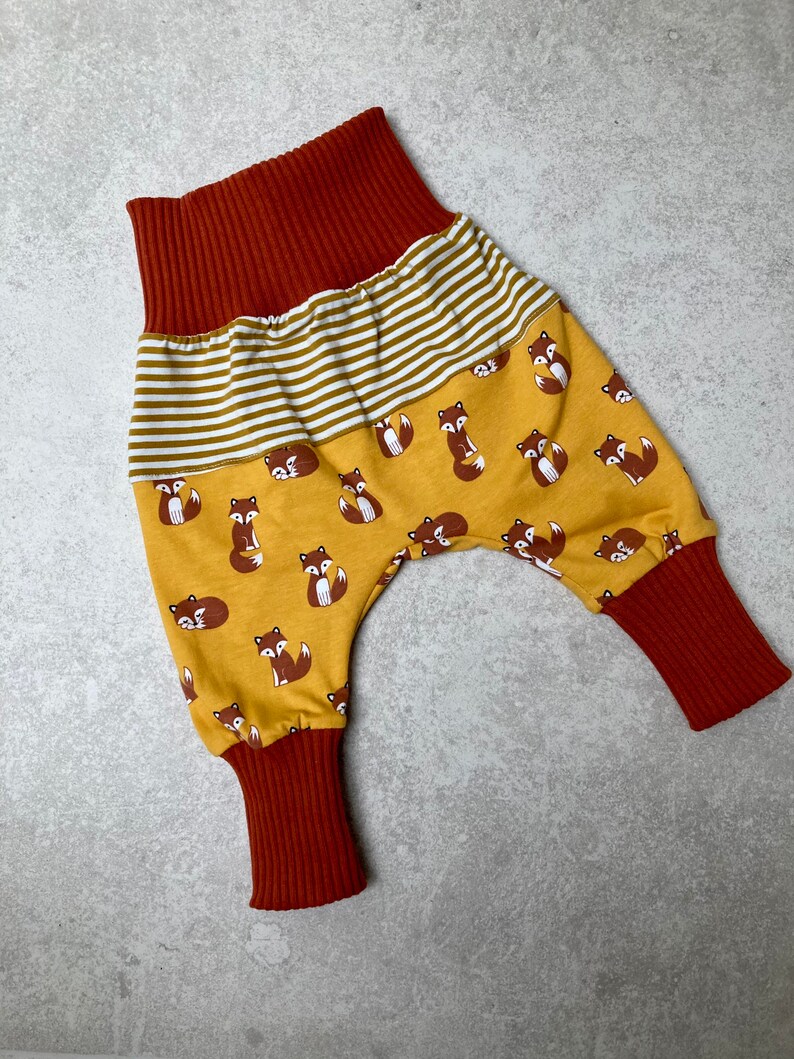 May include: Mustard yellow baby pants with a fox print and red ribbed cuffs. The pants have a white and mustard yellow striped waistband.
