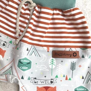 May include: Gray and white striped fabric with a brown stripe. The fabric has a green waistband and a drawstring. The fabric has a pattern of foxes, mountains, trees, and stars. The fabric has the text "Summer Camp" and "Be Wild" printed on it. The fabric has a brown label with the text "Stoffschirurg" printed on it.