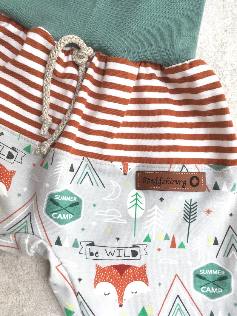 May include: Gray and white striped fabric with a brown stripe. The fabric has a green waistband and a drawstring. The fabric has a pattern of foxes, mountains, trees, and stars. The fabric has the text "Summer Camp" and "Be Wild" printed on it. The fabric has a brown label with the text "Stoffschirurg" printed on it.