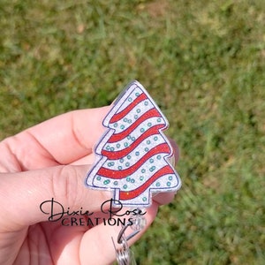 May include: A silver glitter Christmas tree with red and green stripes and white dots. The tree is attached to a clear plastic badge holder with a silver clip.