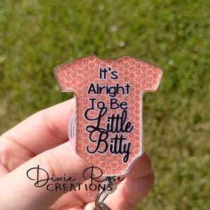 May include: A pink glitter baby onesie with the text "It's Alright To Be Little Bitty" on a clear plastic badge holder.
