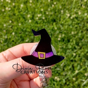 May include: A black witch hat with a purple glitter band and gold trim, attached to a clear retractable badge holder. The text "Dixie Rose Creations" is visible below the hat.