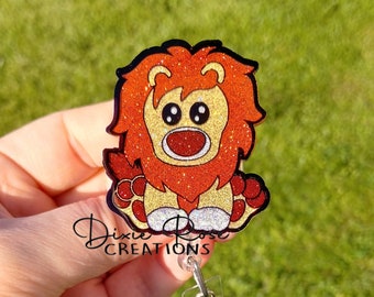 Könnte beinhalten: Ein glitzernder orangefarbener und gelber Löwen-Cartoon-Charakter-Badge-Rollenhalter mit einem silbernen einziehbaren Clip.  Dixie Rose Creations ist auf der Unterseite des Badge-Rollenhalters aufgedruckt.