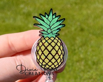 Könnte beinhalten: Ein durchsichtiger Plastik-Badgehalter mit einem Glitzer-Ananas-Design. Die Ananas ist gelb mit grünem Oberteil und schwarzem Umriss. Der Badgehalter hat einen silbernen Clip und eine einziehbare Schnur.