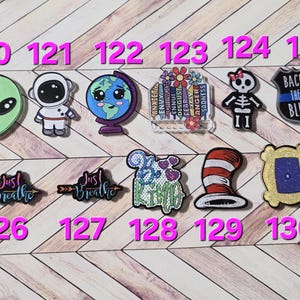 May include: A collection of colorful, cartoon-style charms. Designs include an alien head, astronaut, globe, books with emotions, skeleton, police badge, and text that says "Just Breathe" and "Be Kind".
