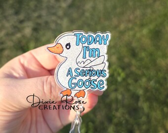 Könnte beinhalten: Ein Abzeichen mit einem Cartoon-Gans-Design. Die Gans ist weiß mit orangefarbenen Füßen und Schnabel, und der Text "Today I'm A Serious Goose" ist in Blau. Das Abzeichen hat einen transparenten Kunststoffclip.