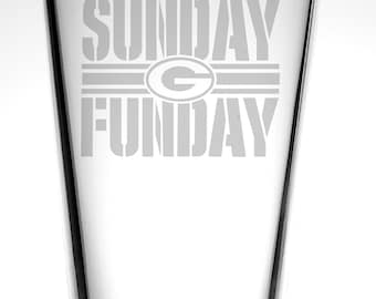 Op de afbeelding: Een helder glazen bierglas met de tekst "SUNDAY FUNDAY" gegraveerd op de voorkant. De tekst is in een vet, bloklettertype. Een groen "G" logo is gecentreerd boven de tekst.