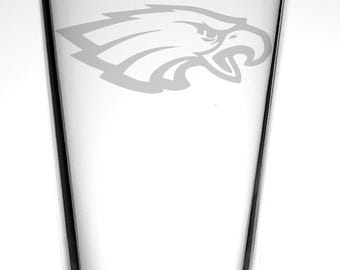 May include: A clear pint glass with a white etched Philadelphia Eagles logo. The logo is a stylized eagle head with a sharp beak and detailed feathers. The glass has a slightly tapered shape and a smooth, transparent surface.