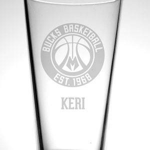May include: A clear glass pint glass with a white etched design of the Milwaukee Bucks basketball team logo. The logo includes the words "Bucks Basketball" and "Est. 1968". The name "Keri" is also etched on the glass.