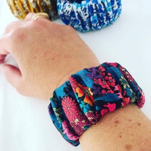 May include: A colorful floral fabric bracelet with pink, blue, and yellow flowers on a dark blue background. The bracelet is made of a soft, stretchy fabric and is perfect for adding a touch of color to any outfit.