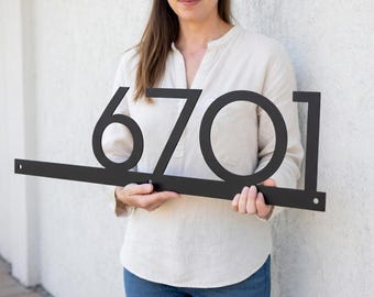 Puede incluir: Un letrero de dirección de metal negro que muestra los números 6701. Los números están en una fuente moderna y están conectados por una barra horizontal. El letrero lo sostiene una persona frente a una pared blanca.