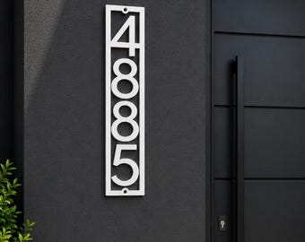 Puede incluir: Una placa de número de casa rectangular vertical que presenta los dígitos blancos 4, 8, 8 y 5 apilados dentro de un marco limpio. El letrero está montado en una pared gris oscuro junto a una puerta negra, mostrando los números en una fuente sans-serif.