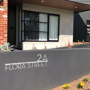 May include: A modern house with a gray exterior and a black door. The house has a black metal sign that reads "2A FLORA STREET".