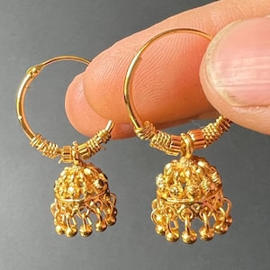 May include: A pair of gold-coloured hoop earrings. Each earring has a detailed, bell-shaped charm with dangling small spheres. The hoops are held by a hand against a grey background. The earrings have a bright, polished finish.