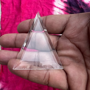 May include: A clear glass pyramid with a pointed top and a flat base. The pyramid is held in a hand.