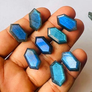 Könnte beinhalten: Eine Sammlung facettierter Labradorit-Edelsteine in verschiedenen Blau- und Türkistönen. Jeder Stein hat eine einzigartige sechseckige Form mit polierten Oberflächen, die irisierende Blitze zeigen. Die Edelsteine werden in einer Hand gehalten.