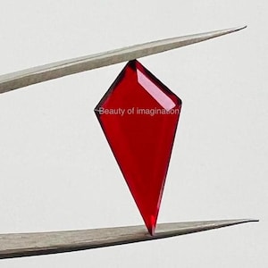 May include: A bright red, faceted, teardrop-shaped glass gem held by tweezers. The gem is clear and has a slight sparkle.