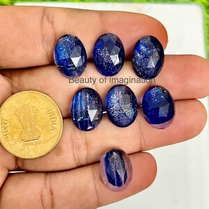 Peut inclure: Sept cabochons ovales en pierre précieuse bleue avec des inclusions de paillettes argentées. Les cabochons sont présentés sur une main.