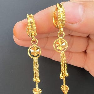 Op de afbeelding: Een paar goudkleurige oorbellen. Elke oorbel heeft een ring, een cirkelvormige hanger met een sterrenpatroon, een ketting van kleine schakels en een hartvormige bedel. De oorbellen worden tegen een donkere achtergrond getoond.