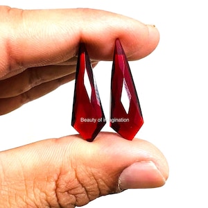 May include: Two red faceted gemstone drops, held between fingers, with a white background.