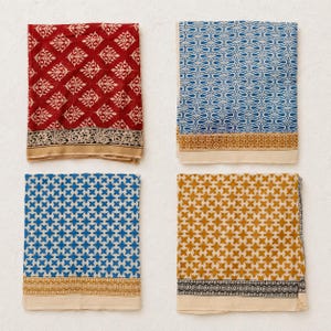 May include: Four square fabric napkins arranged in a grid. Top left is red with a white floral pattern. Top right is blue with a white teardrop pattern. Bottom left is blue with a white cross pattern. Bottom right is gold with a white cross pattern.