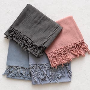 May include: Three rectangular scarves in charcoal grey, dusty rose, and slate blue. Each scarf features a smooth, solid-coloured fabric texture and is finished with long, knotted fringe tassels along the bottom edges, arranged in a layered stack.