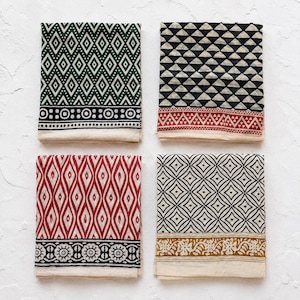 May include: Four folded square fabric napkins featuring distinct block-printed patterns. One has green diamonds, one black triangles with a red border, one red elongated diamonds, and one black geometric shapes with a mustard floral border.