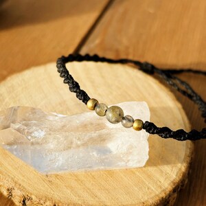 Puede incluir: Una pulsera de macramé negra con cierre de nudo corredizo. La pulsera presenta tres cuentas grises y doradas. La pulsera se exhibe sobre una rodaja de madera con un cristal transparente.