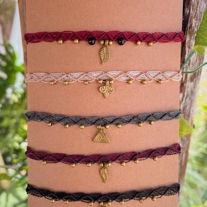 Puede incluir: Cinco gargantillas de macramé hechas a mano en varios colores: burdeos, beige, gris, rojo oscuro y negro. Cada gargantilla presenta un colgante único y pequeñas cuentas doradas. Los colgantes incluyen diseños de hojas y geométricos.