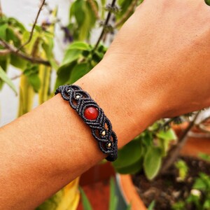 Peut inclure: Un bracelet en macramé noir avec une perle de cornaline rouge. Le bracelet est noué et comporte des accents dorés.