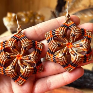 Peut inclure: Une paire de boucles d'oreilles en macramé en forme d'étoile avec un motif géométrique dans des tons de beige, brun foncé et orange vif. Les boucles d'oreilles sont faites de cordon noué et incluent des crochets en métal argenté.