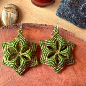 Peut inclure: Une paire de boucles d'oreilles en macramé en forme d'étoile, réalisées avec un cordon vert olive et des accents roses discrets. Chaque boucle présente un motif géométrique à six branches avec une ouverture centrale et un crochet en métal argenté.