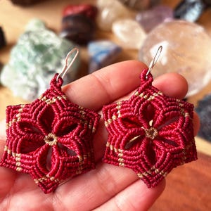 May include: A pair of macrame earrings featuring a six-pointed star or flower shape. The design is crafted from deep red and tan cord, woven into a geometric pattern with a central opening. Each earring is attached to a silver-toned metal hook.