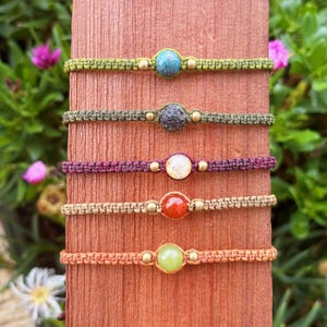 Puede incluir: Cinco pulseras de macramé con piedras de diferentes colores. Las pulseras son en tonos verdes, morados, beige y naranjas. Cada pulsera tiene una piedra central y cuentas doradas.