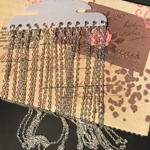 May include: A close-up of a display of silver chain necklaces. The chains are hanging from a white plastic display card with a hanger. The chains are all different lengths and thicknesses.