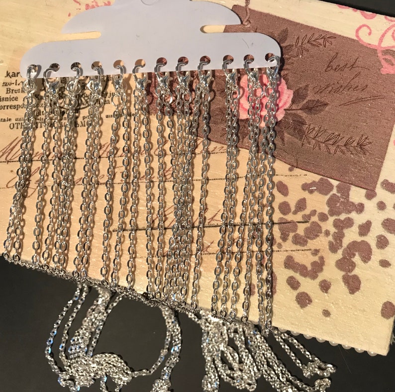 May include: A close-up of a display of silver chain necklaces. The chains are hanging from a white plastic display card with a hanger. The chains are all different lengths and thicknesses.