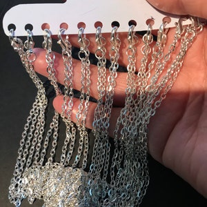 May include: A close-up of a silver chain necklace on a white display card. The chain is made of small, linked oval shapes. There are multiple chains on the card, all of the same style.