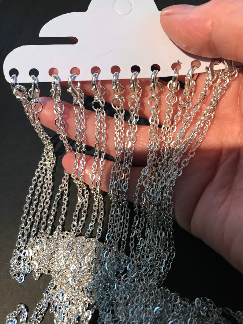 May include: A close-up of a silver chain necklace on a white display card. The chain is made of small, linked oval shapes. There are multiple chains on the card, all of the same style.