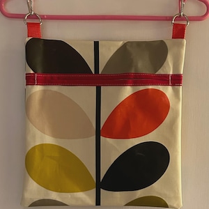 May include: A cream-colored tote bag with a colorful leaf pattern in shades of brown, orange, yellow, and black. The bag has a red strap and is suspended from a pink hanger with silver clips.